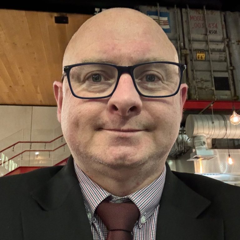 Mark Smart Mark in a suit and tie, wearing glasses, in a restaurant setting.